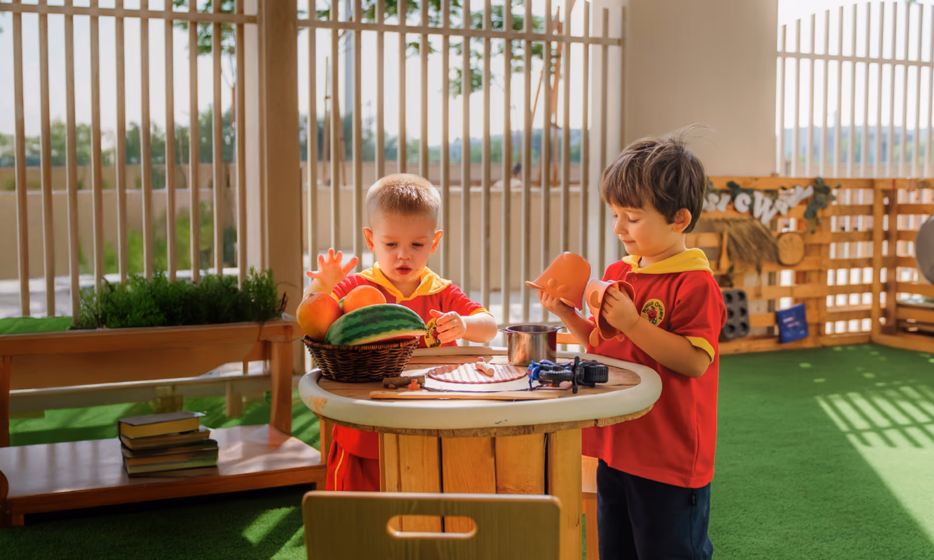 Two young boys in red and yellow uniforms playing with toy kitchen utensils and a basket of play fruit at a round wooden table in a sunlit indoor play area.