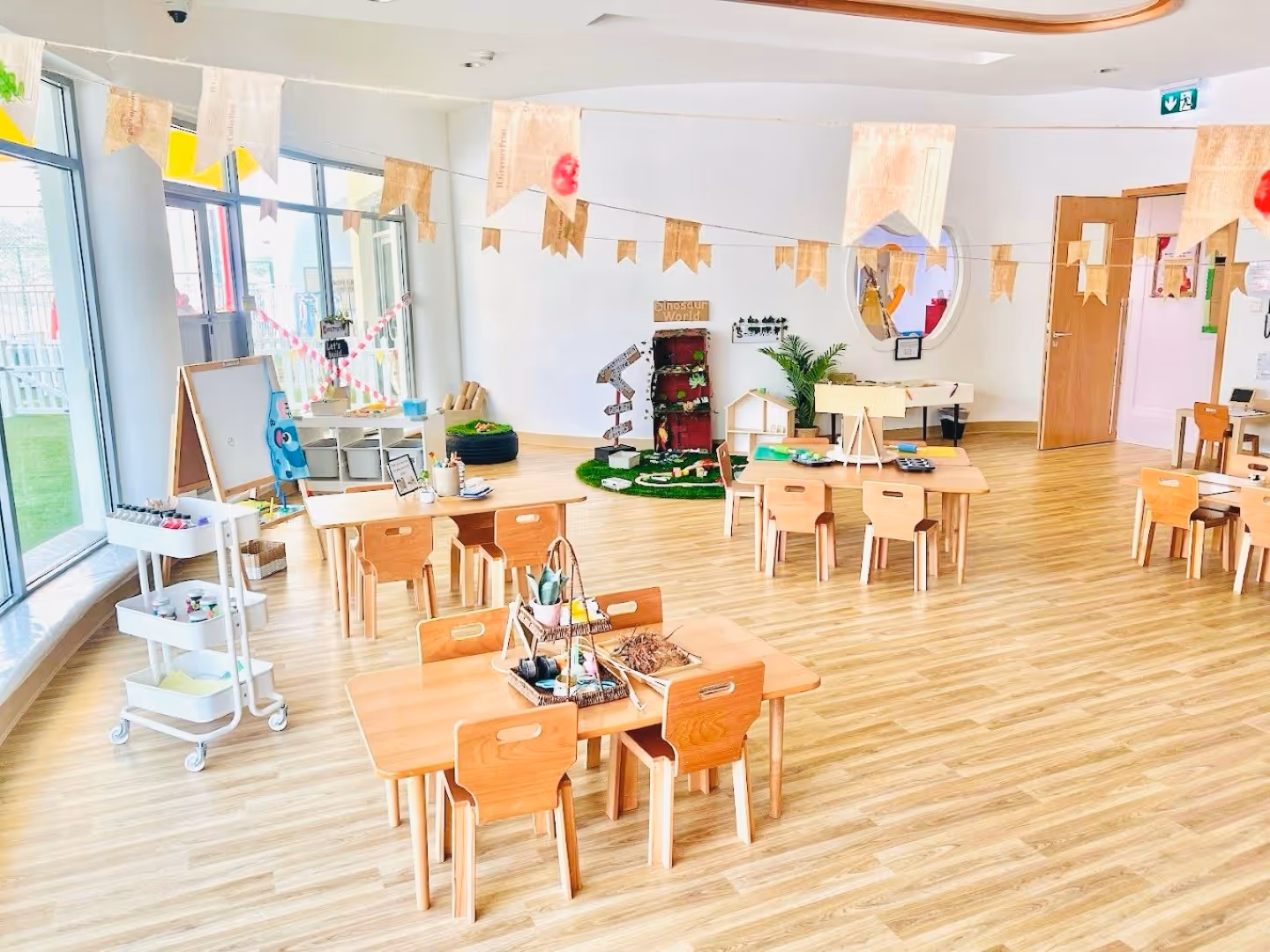 Bright and spacious preschool classroom with wooden tables and chairs, art supplies, and educational play areas.