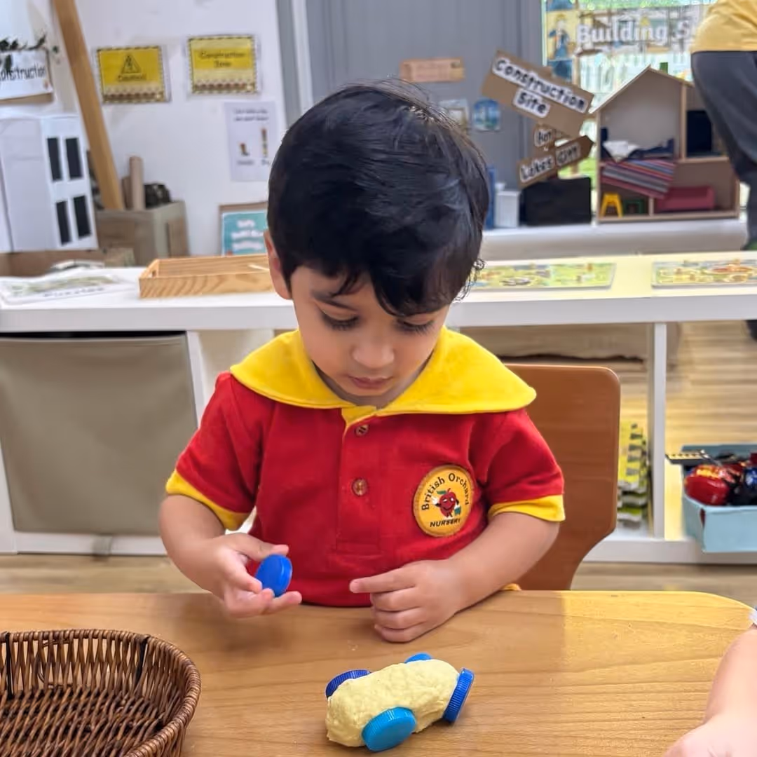 Young child in a red and yellow British Orchard Nursery uniform playing with a dough vehicle with blue bottle caps as wheels at a wooden table.