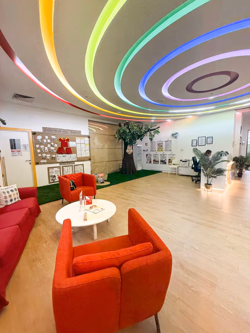 Bright indoor lounge with red chairs and sofa, a white round table, a bulletin board, a tree decoration, and colorful ceiling lights.