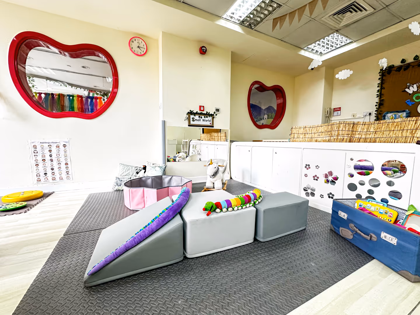 Bright and colorful indoor children's play area with soft foam shapes, toys, and apple-shaped mirrors on the walls.