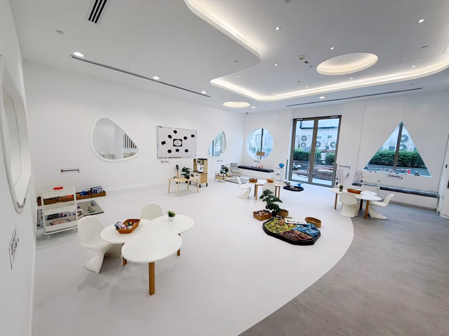 Bright modern classroom with white walls, tables and chairs, categorized activity stations, and large geometric windows.