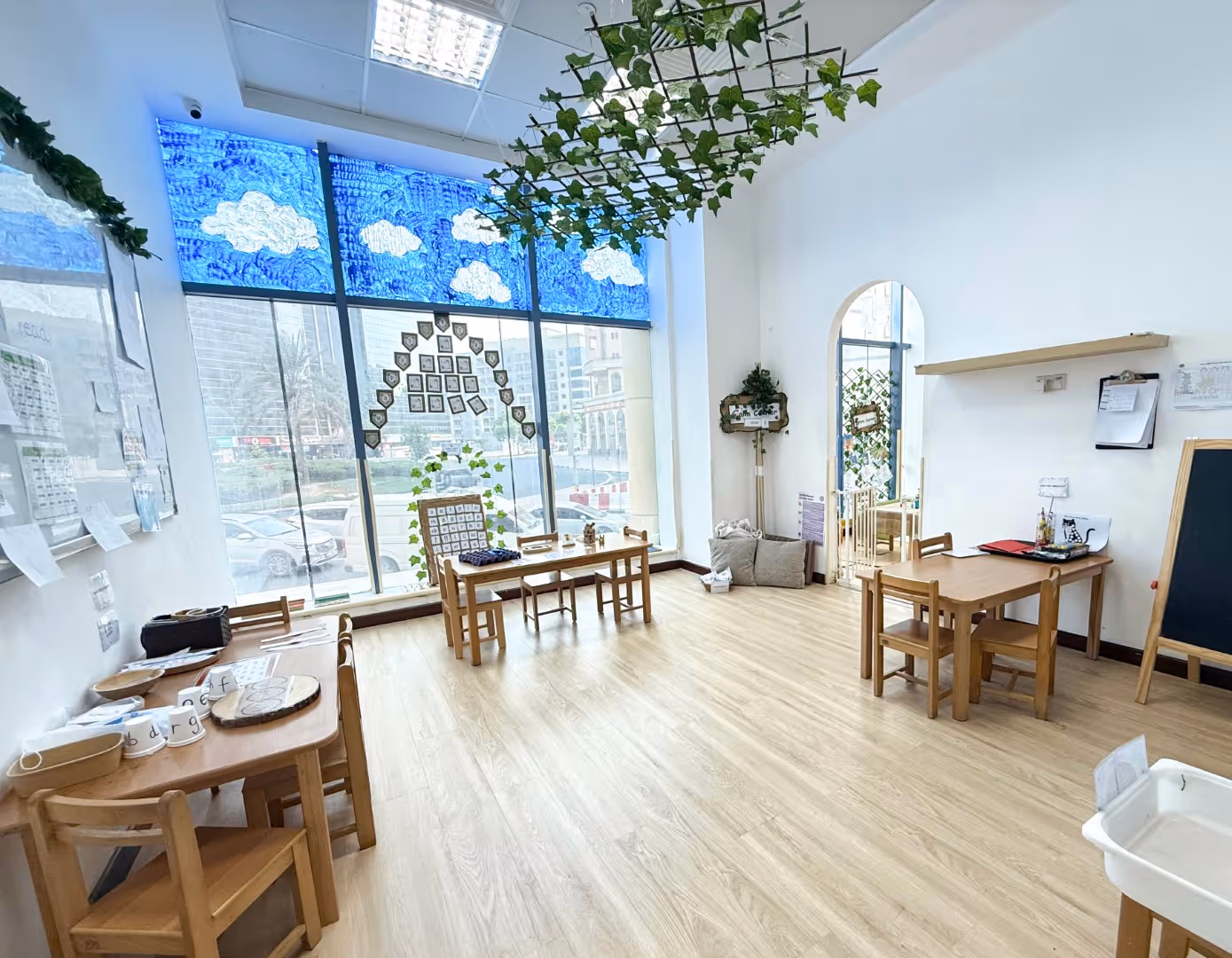 Bright classroom with wooden tables and chairs, a large window decorated with blue sky and cloud patterns, and green vines hanging from the ceiling.