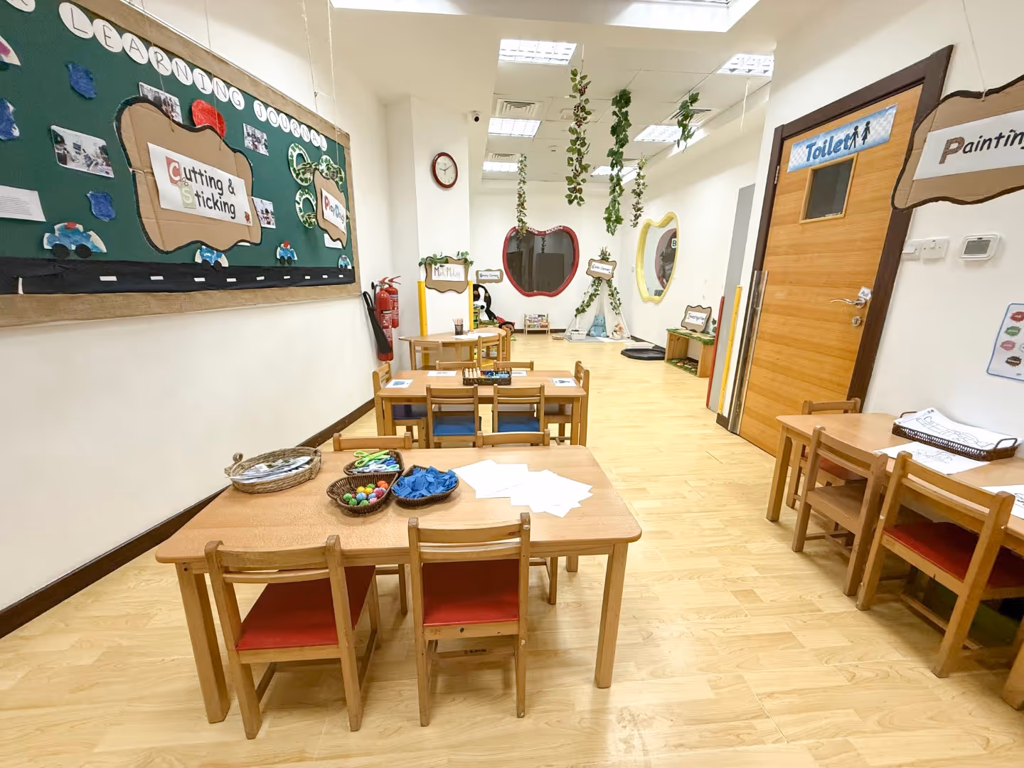 Bright and organized classroom with wooden tables and chairs, children's art supplies, hanging plants, and educational wall displays.