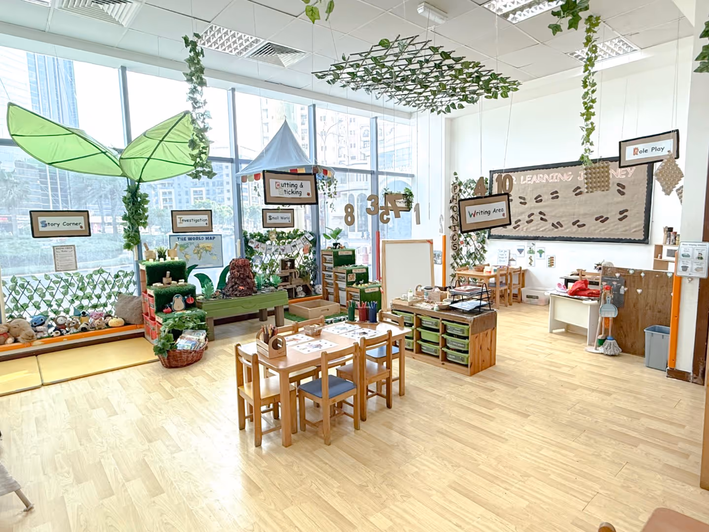 Bright and colorful early childhood classroom with various learning stations, small wooden tables and chairs, educational posters, and hanging greenery decorations.
