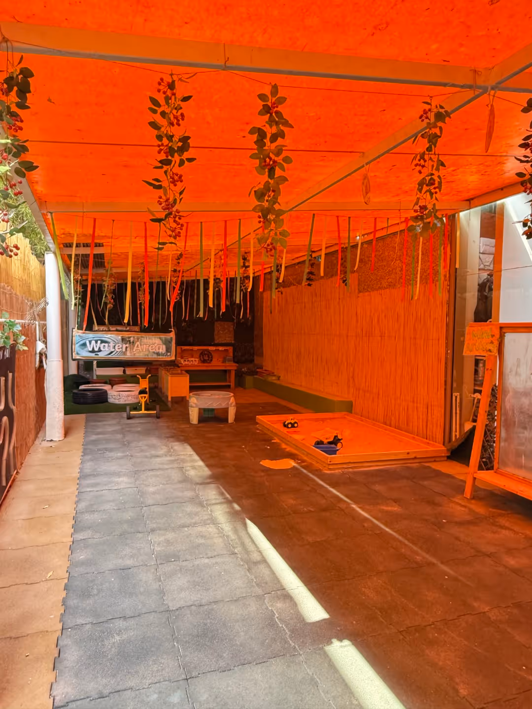 Covered outdoor play area with hanging decorations, play tables, and children’s toys under an orange canopy.