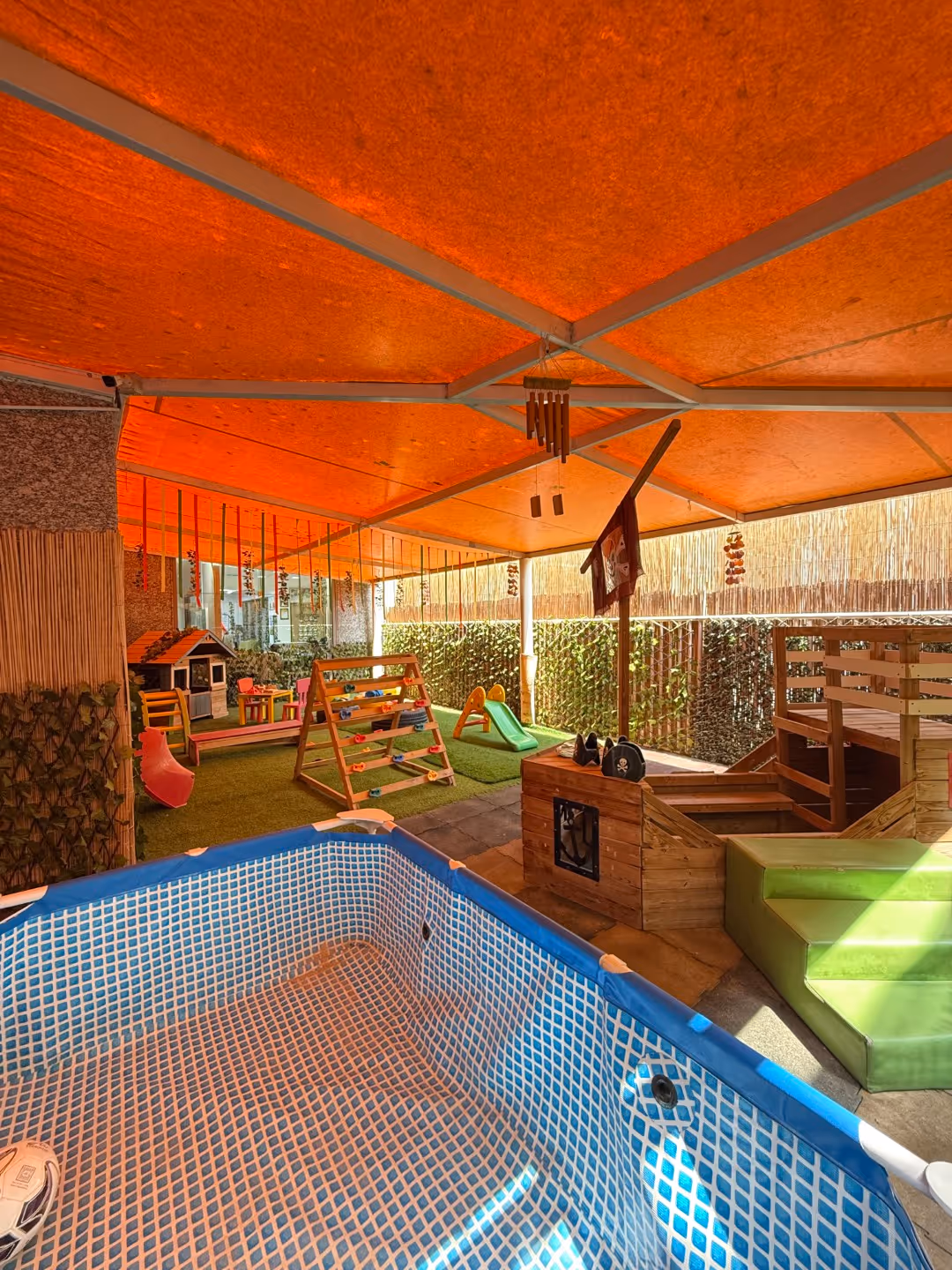 Indoor children's play area with a small empty pool, wooden play structures including a climbing frame and slide, under an orange canopy.