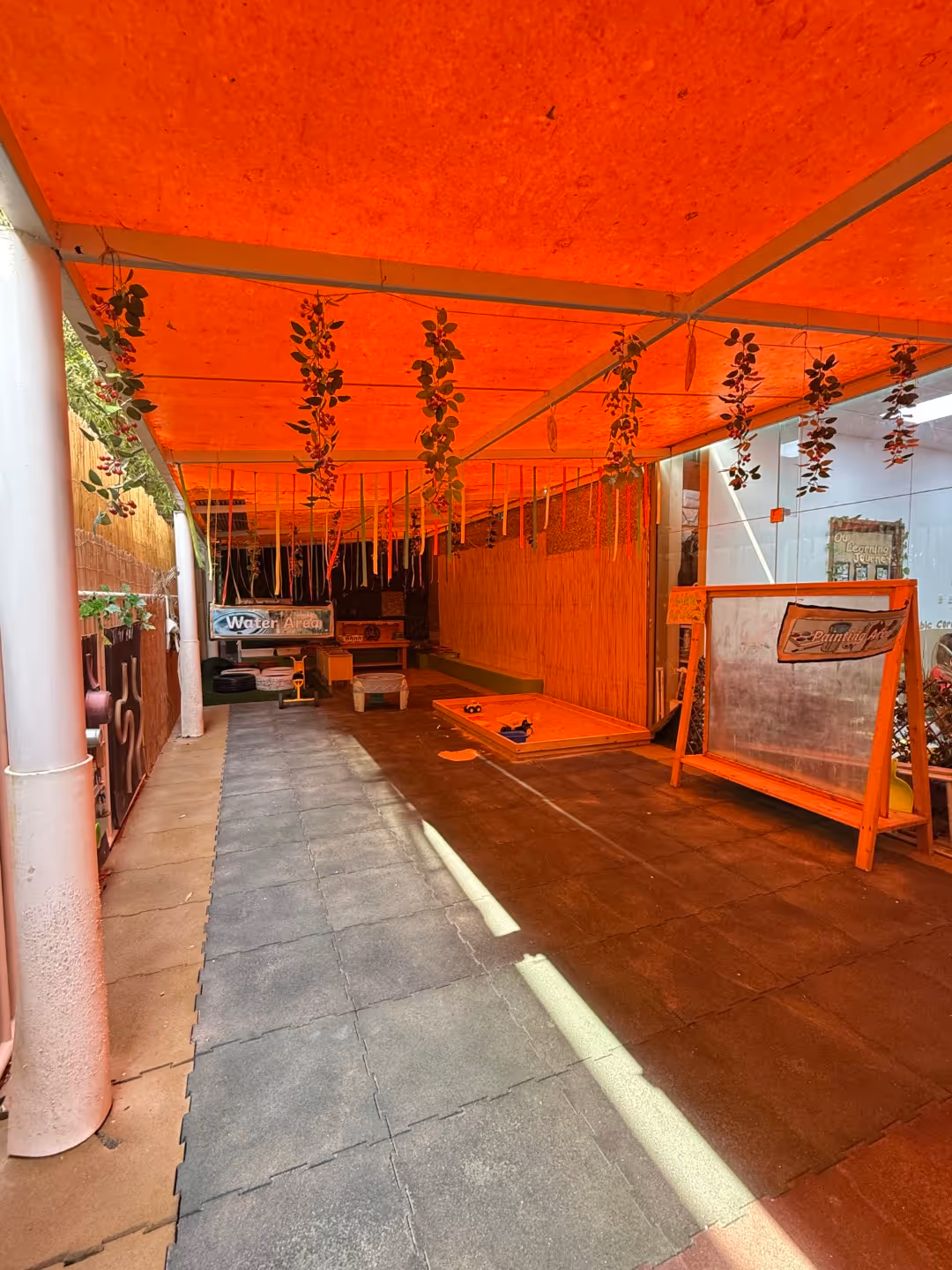 Covered play area with hanging plants, foam floor tiles, a sandbox, and a clear board labeled 'Painting Area'.