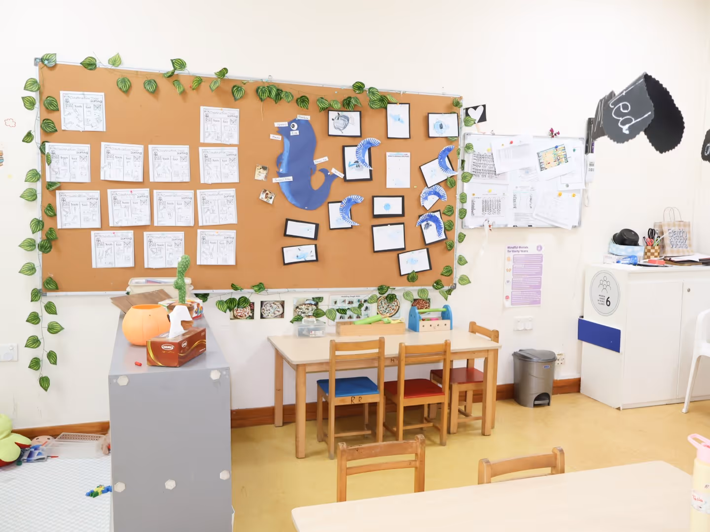 Classroom corner with children's table and chairs, bulletin board decorated with papers, blue seahorse crafts, and climbing plant decorations.