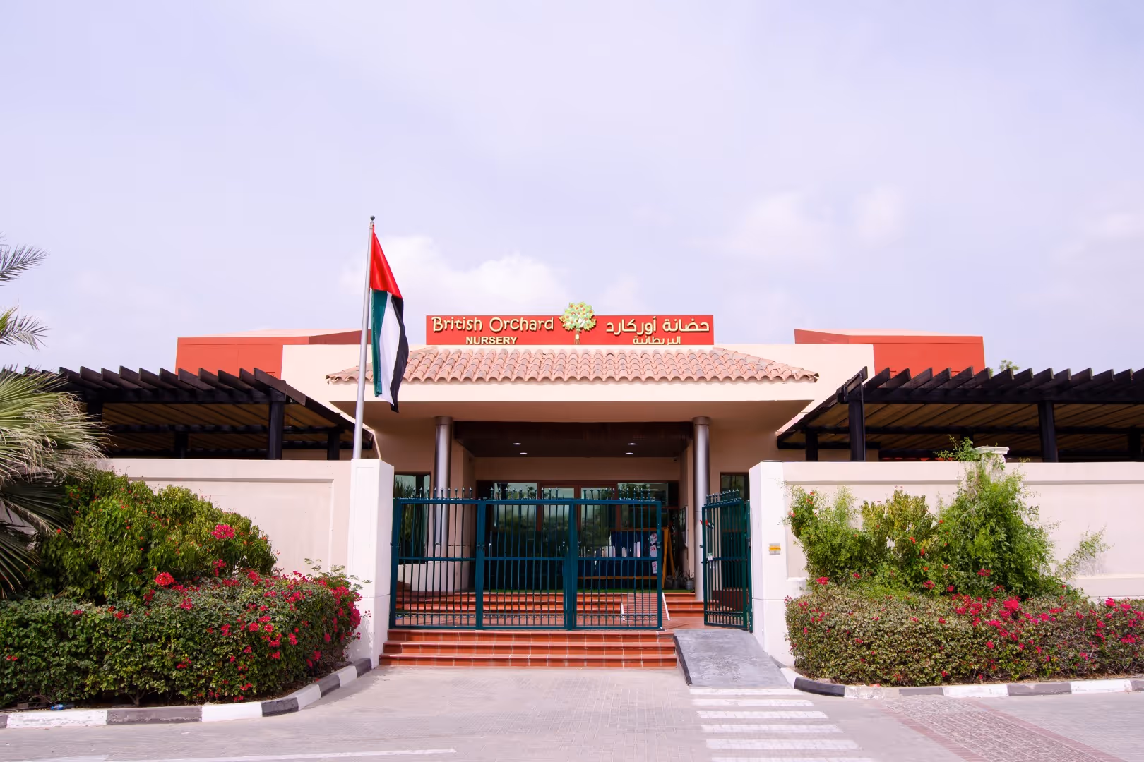 Entrance of British Orchard Nursery with green gate, UAE flag on a pole, and surrounding greenery with red flowers.