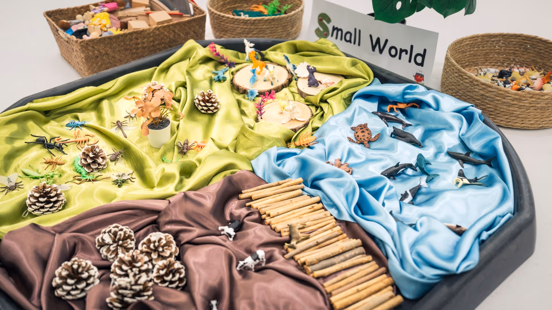 Sensory play tray divided into green, blue, and brown sections with toy jungle animals, sea creatures, pine cones, and wooden sticks labeled 'Small World.'