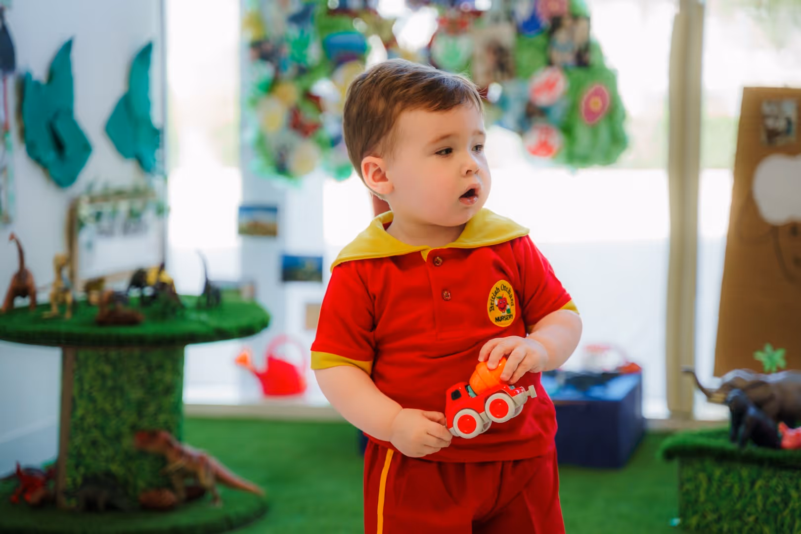 Toddler in a red and yellow nursery uniform holding a red toy truck with an orange object indoors.