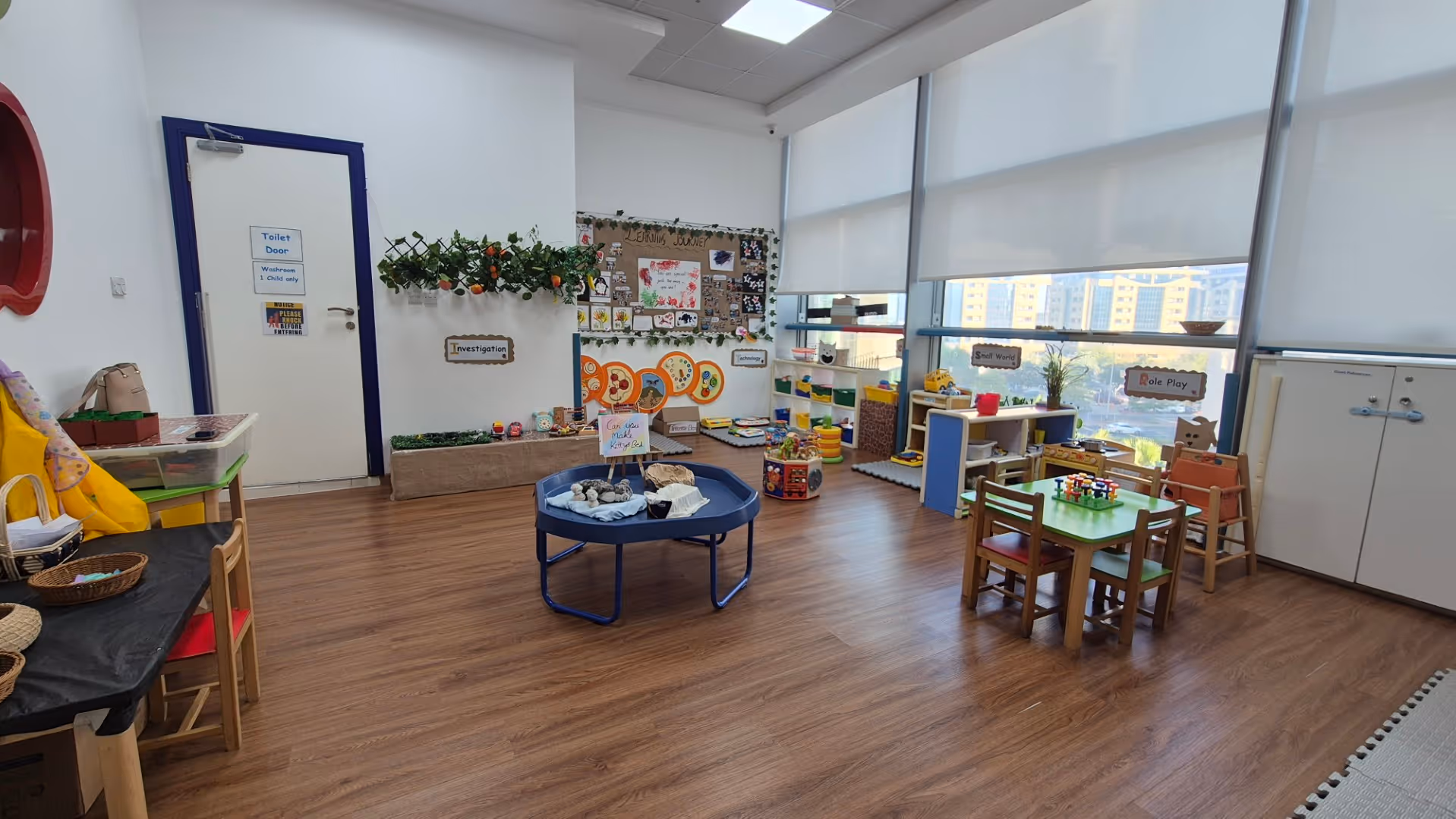 Bright children’s classroom with wooden flooring, small tables and chairs, educational toys, and wall displays including 'Investigation' and 'Role Play'.
