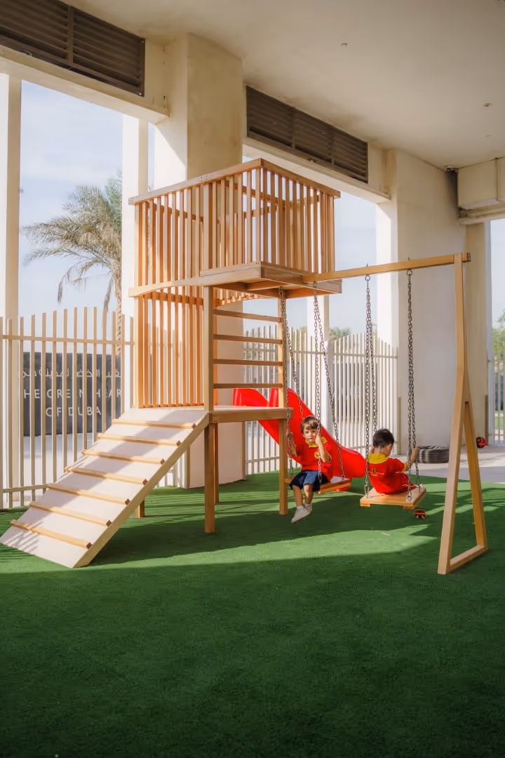 Two young children in red shirts playing on wooden swings attached to a play structure with a red slide in a covered playground with green artificial grass.