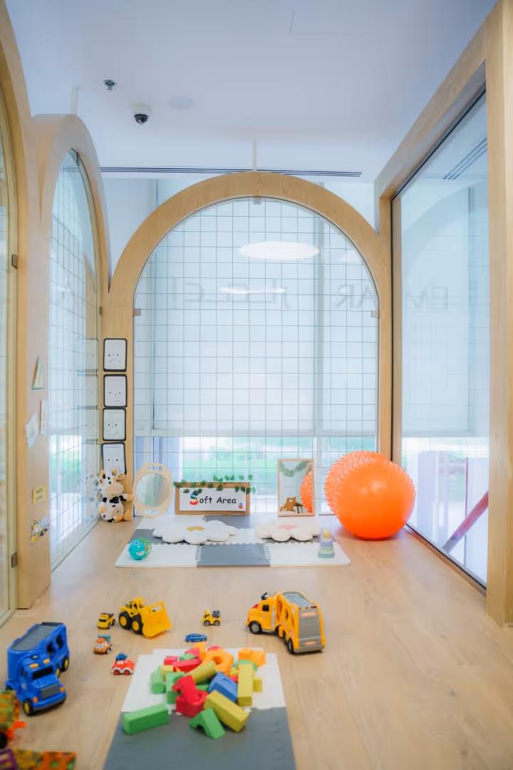 Bright children's play area with wooden floors, colorful toy blocks, toy vehicles, stuffed animals, a mirror, and an orange exercise ball.