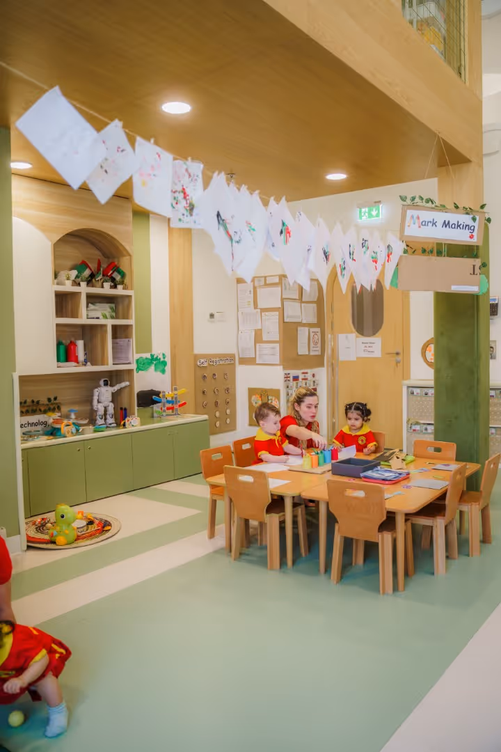 A teacher and two children in red shirts sitting at a wooden table in a classroom decorated with children's artwork and educational toys.