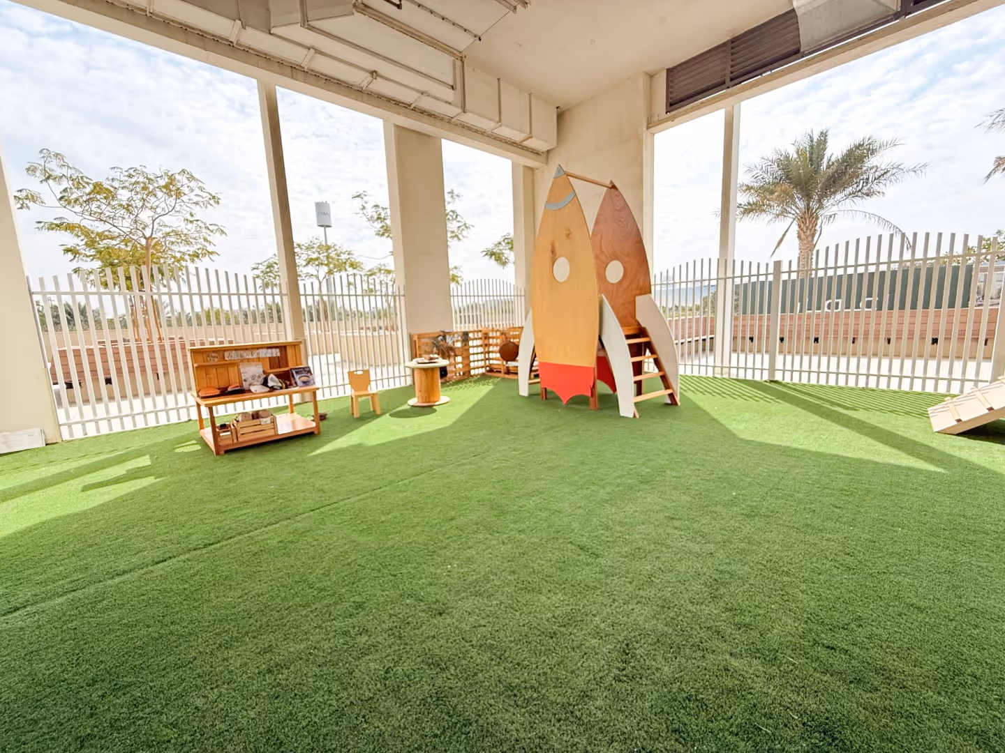 Indoor play area with artificial grass featuring a wooden rocket-shaped play structure and small furniture pieces near large barred windows.