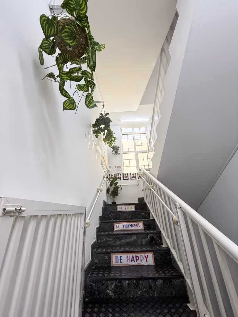 Staircase with black steps featuring motivational signs reading 'BE HAPPY,' 'BE THANKFUL,' and 'LOVE YOU,' with hanging green plants and a large window at the top.