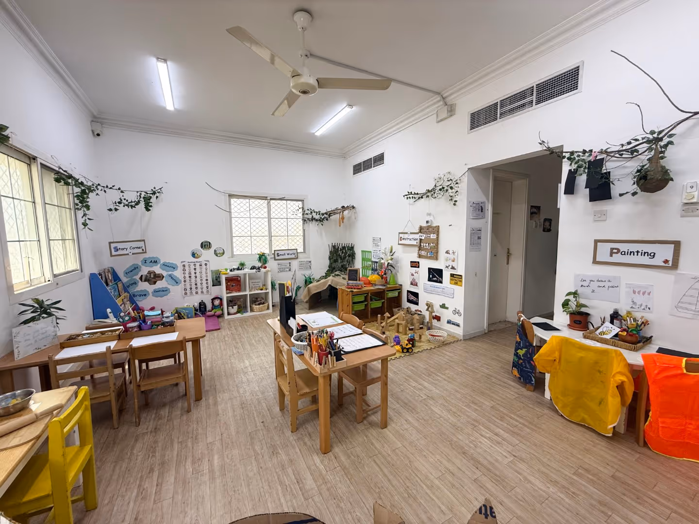 Bright preschool classroom with small wooden tables and chairs, educational posters, and various learning and play areas including story corner and painting station.
