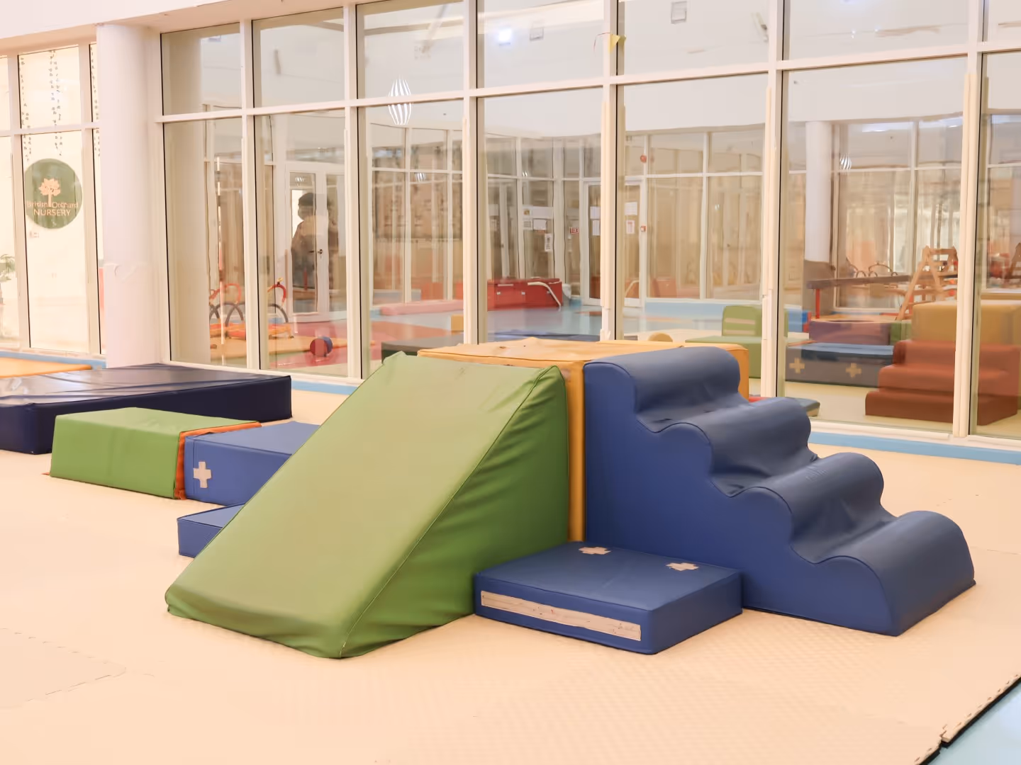 Indoor play area with soft padded climbing steps, a green slide, and cushioned mats near glass windows.