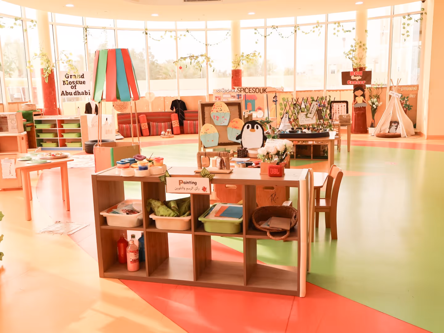 Bright classroom with art supplies, colorful floor, and various creative displays including a penguin craft and a tent.