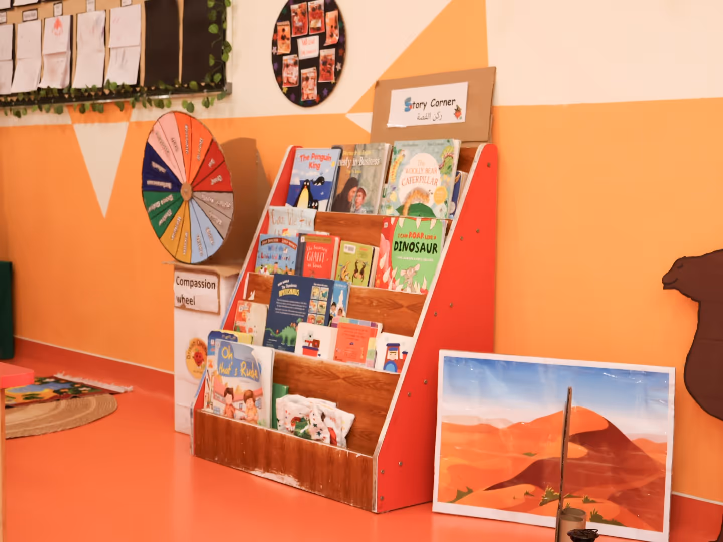 Children's story corner with a wooden red and white bookshelf filled with colorful books and a compassion wheel on the wall.