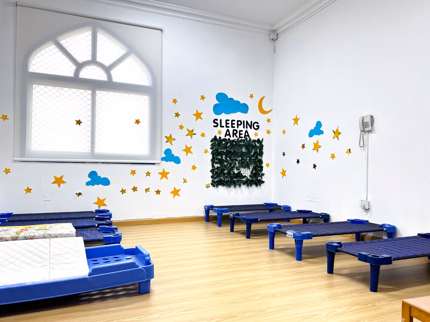 Bright sleeping area with small blue beds arranged in rows on a wooden floor and decorated white walls with stars, clouds, and a crescent moon.