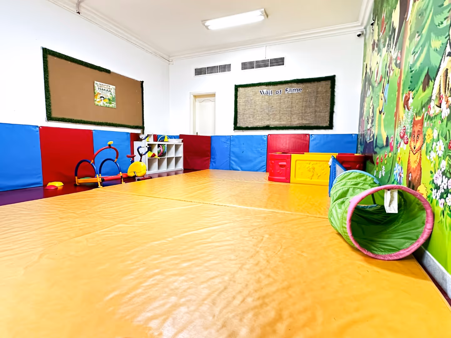 Indoor children's playroom with padded floor mats, colorful toys, storage cubbies, and a wall mural featuring forest animals.