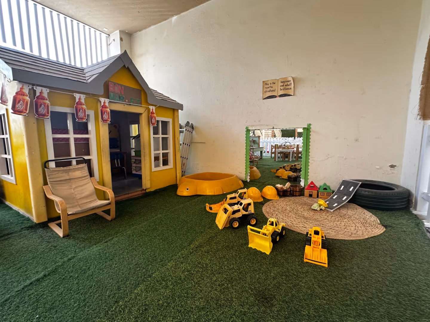 Indoor play area with a yellow playhouse, toy construction vehicles, helmets, a small rug, and a mirror on a wall.