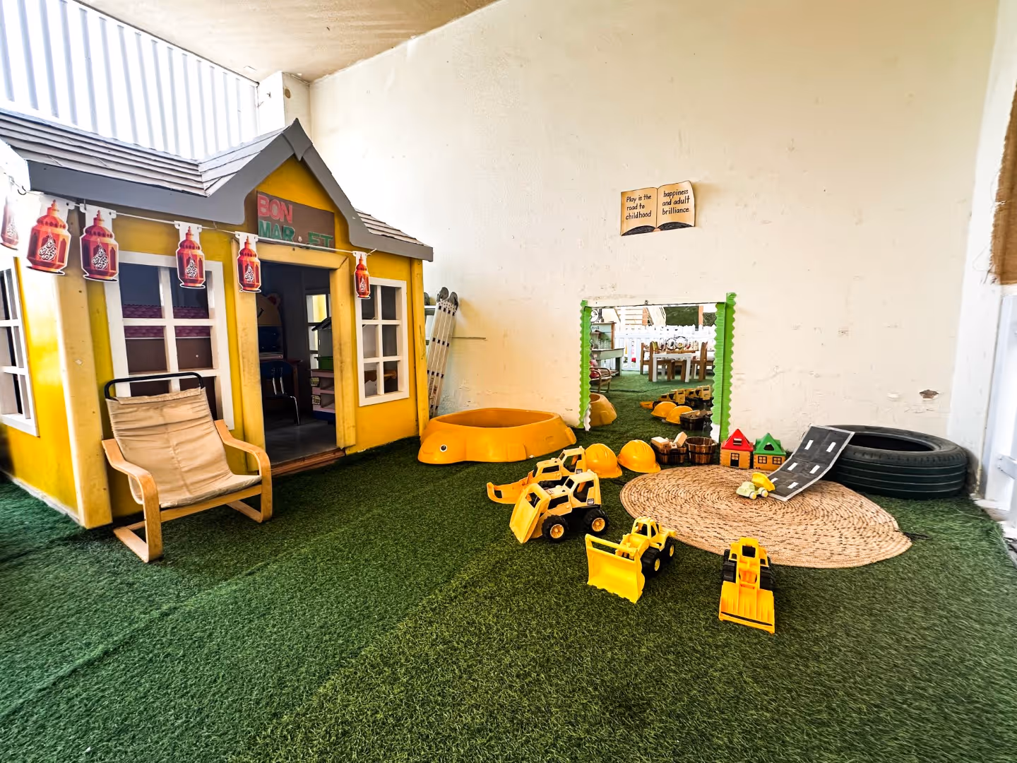 Indoor play area with a yellow playhouse, toy construction vehicles, helmets, a small mirror, and artificial grass flooring.