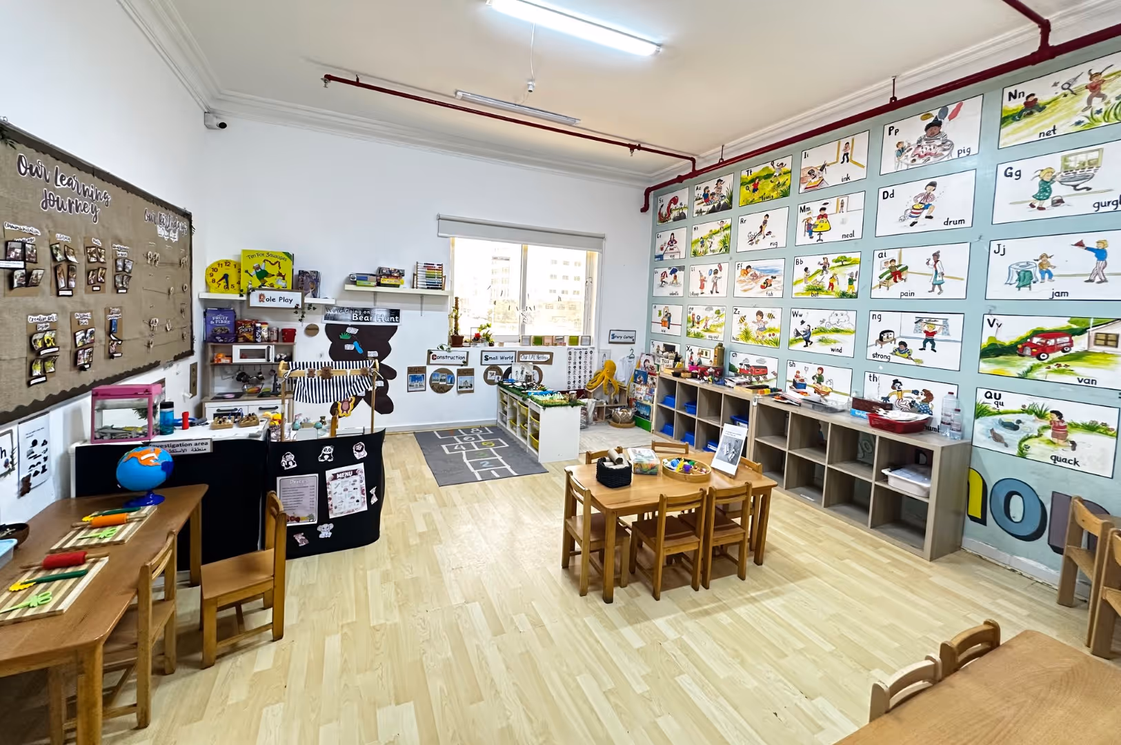 Bright preschool classroom with wooden tables and chairs, alphabet wall posters, educational toys, and activity stations.
