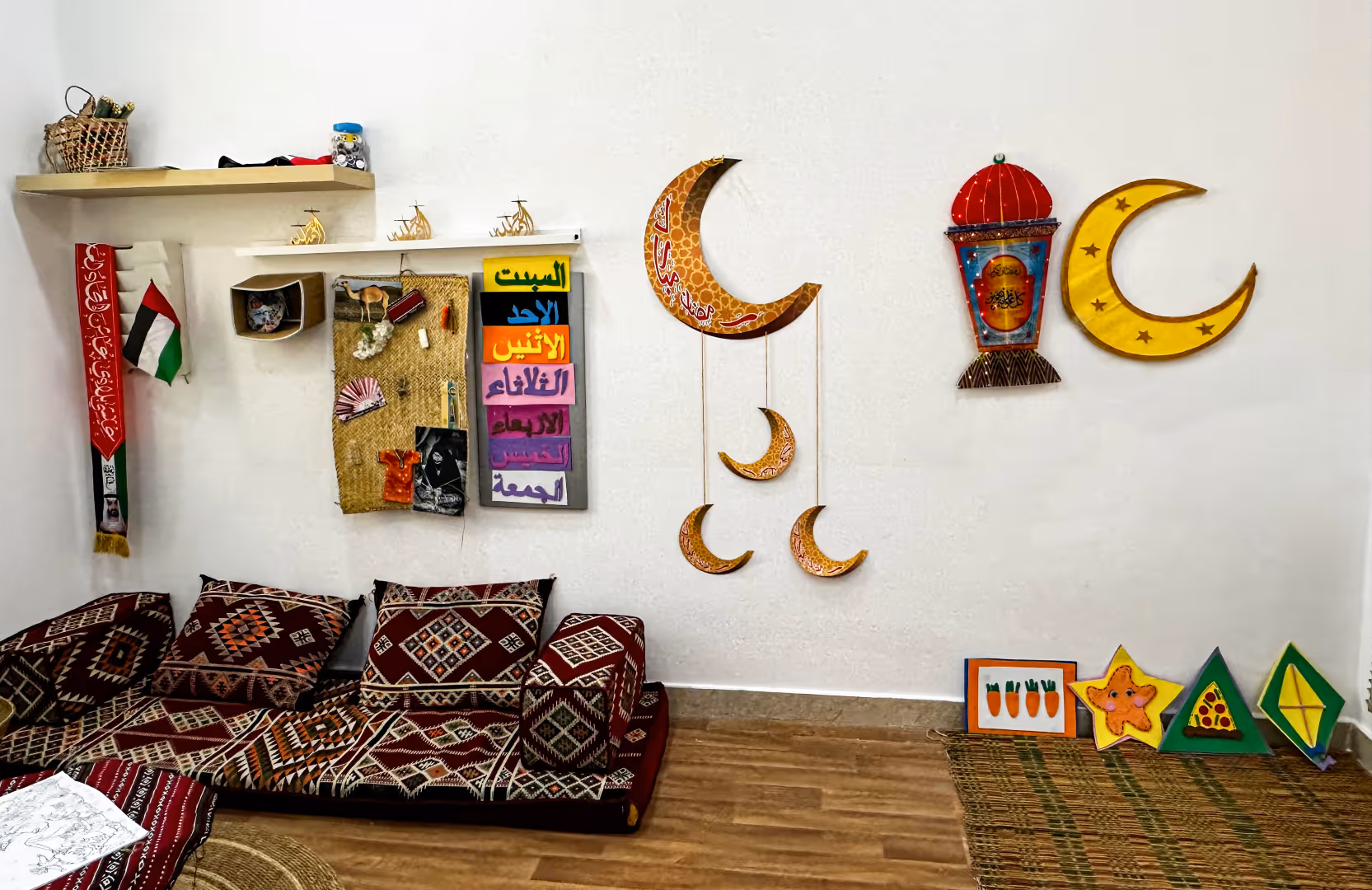 Cozy corner sofa with patterned cushions against a white wall decorated with hanging moons, a lantern, a small UAE flag, colorful block signs with Arabic days of the week, and playful star and kite decorations on a woven mat floor.