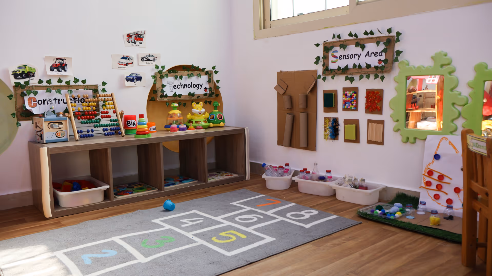 Bright children's playroom corner with educational toys, numbered hopscotch rug, and sensory activity area.
