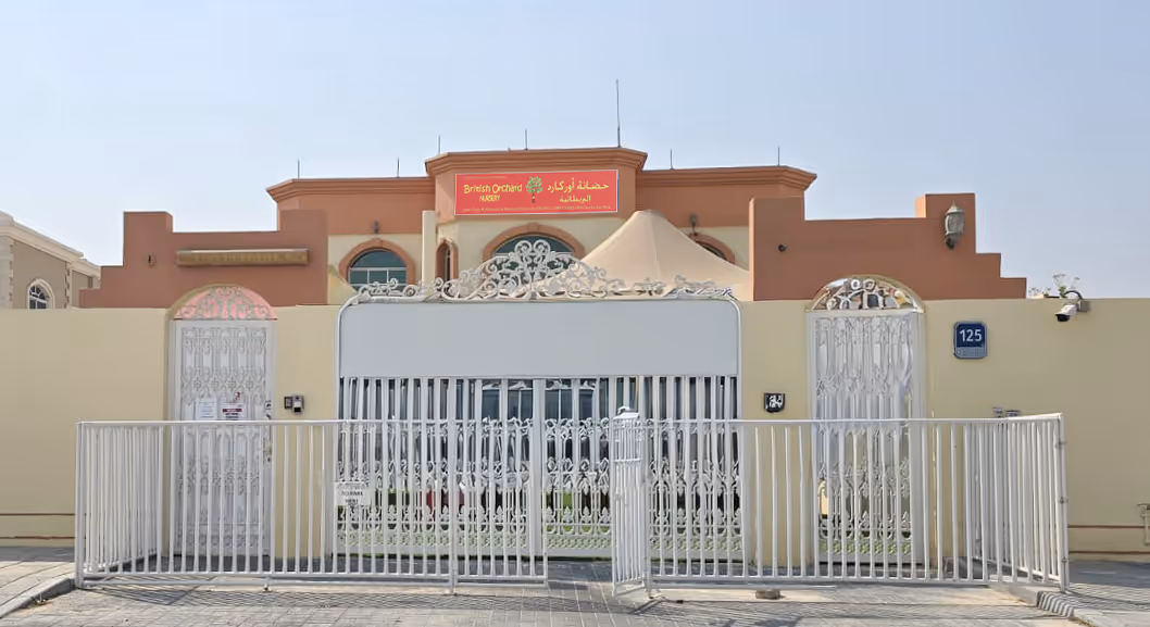 Exterior view of British Orchard Nursery building behind a decorative white gate and fencing with the nursery's sign in red and yellow on top.