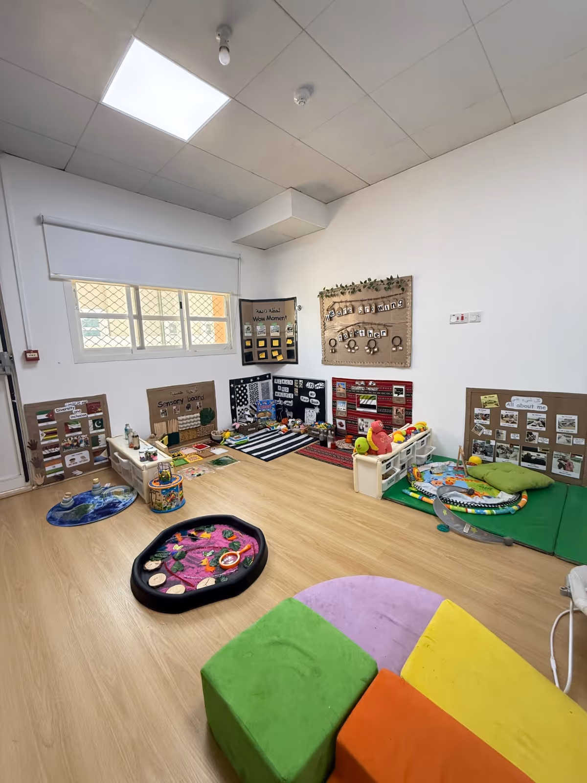 Bright children's playroom with sensory boards, toy storage, colorful seating cubes, and various educational play items on a wooden floor.