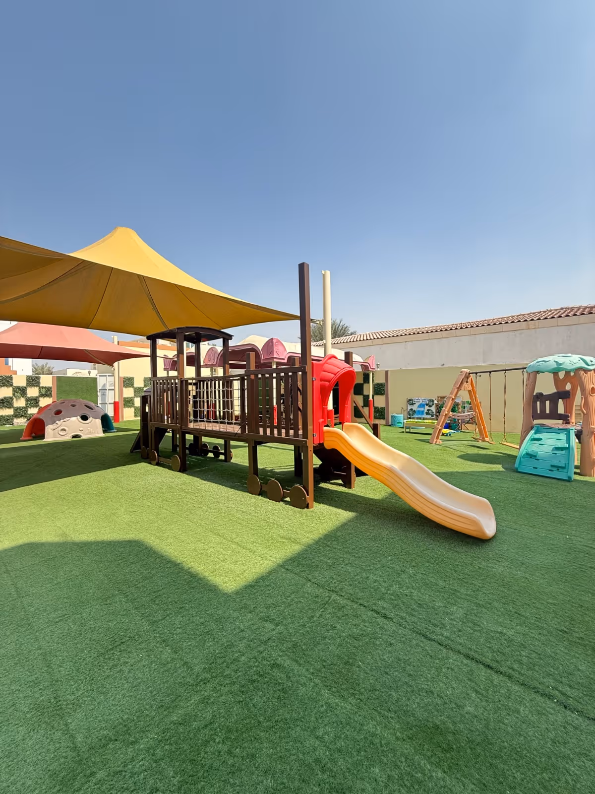 Outdoor children's playground with artificial grass, a yellow slide attached to a brown play structure under a large yellow canopy, swings, and other play equipment.