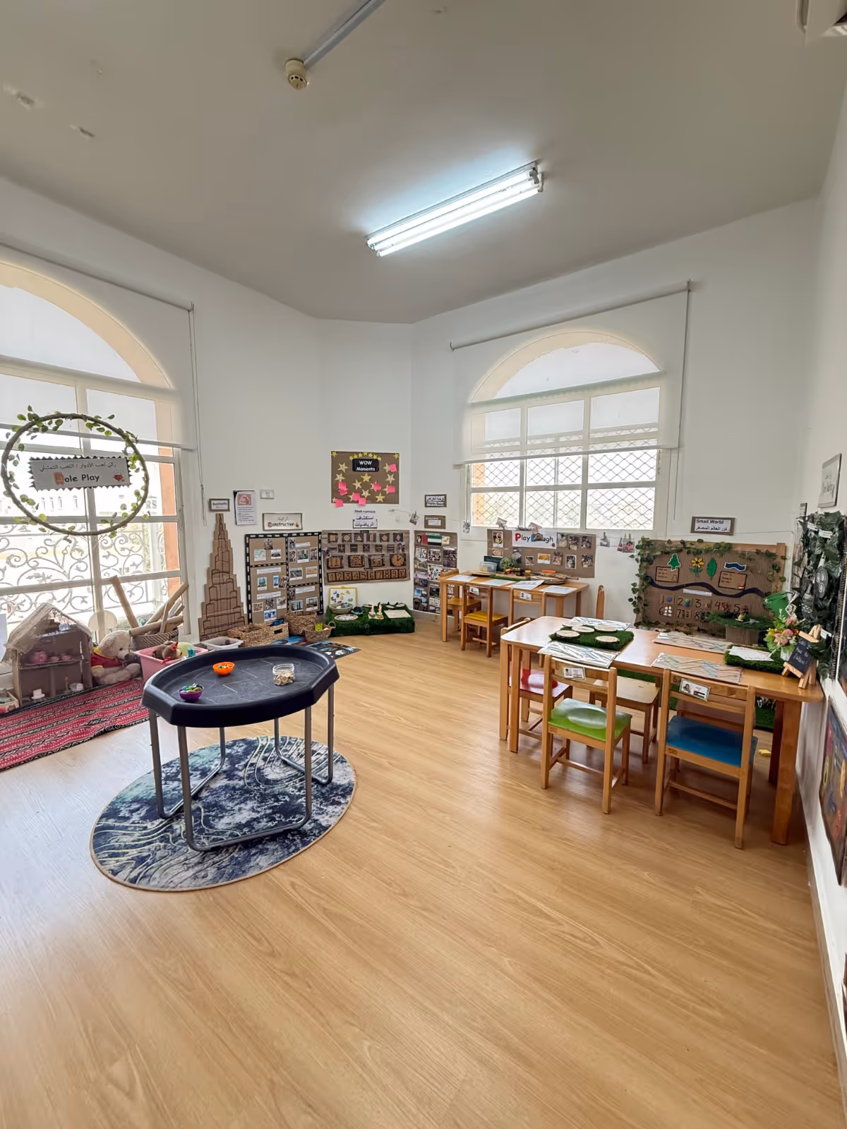 Bright classroom with wooden chairs and tables, educational displays on walls, and a small round activity table on a rug.