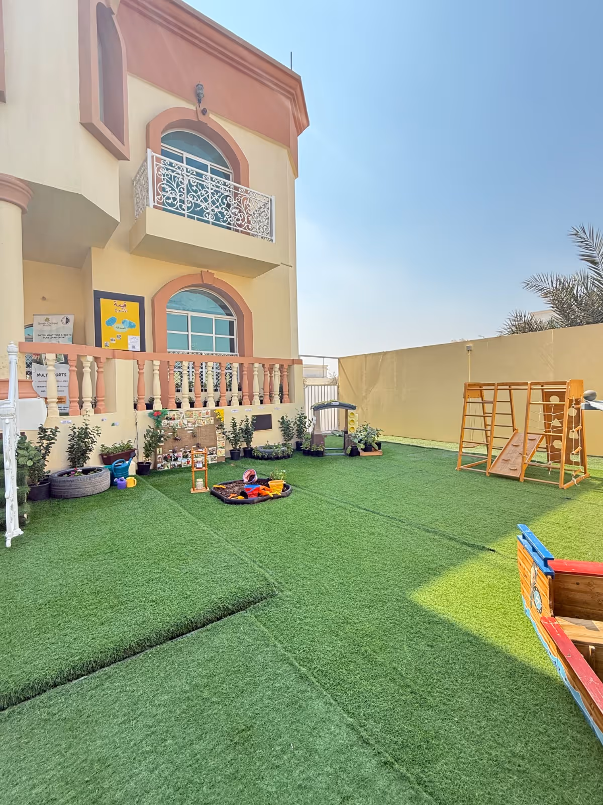 Outdoor children's play area with artificial grass, climbing frame, sandbox with toys, plants in pots, and a wooden play structure shaped like a boat.