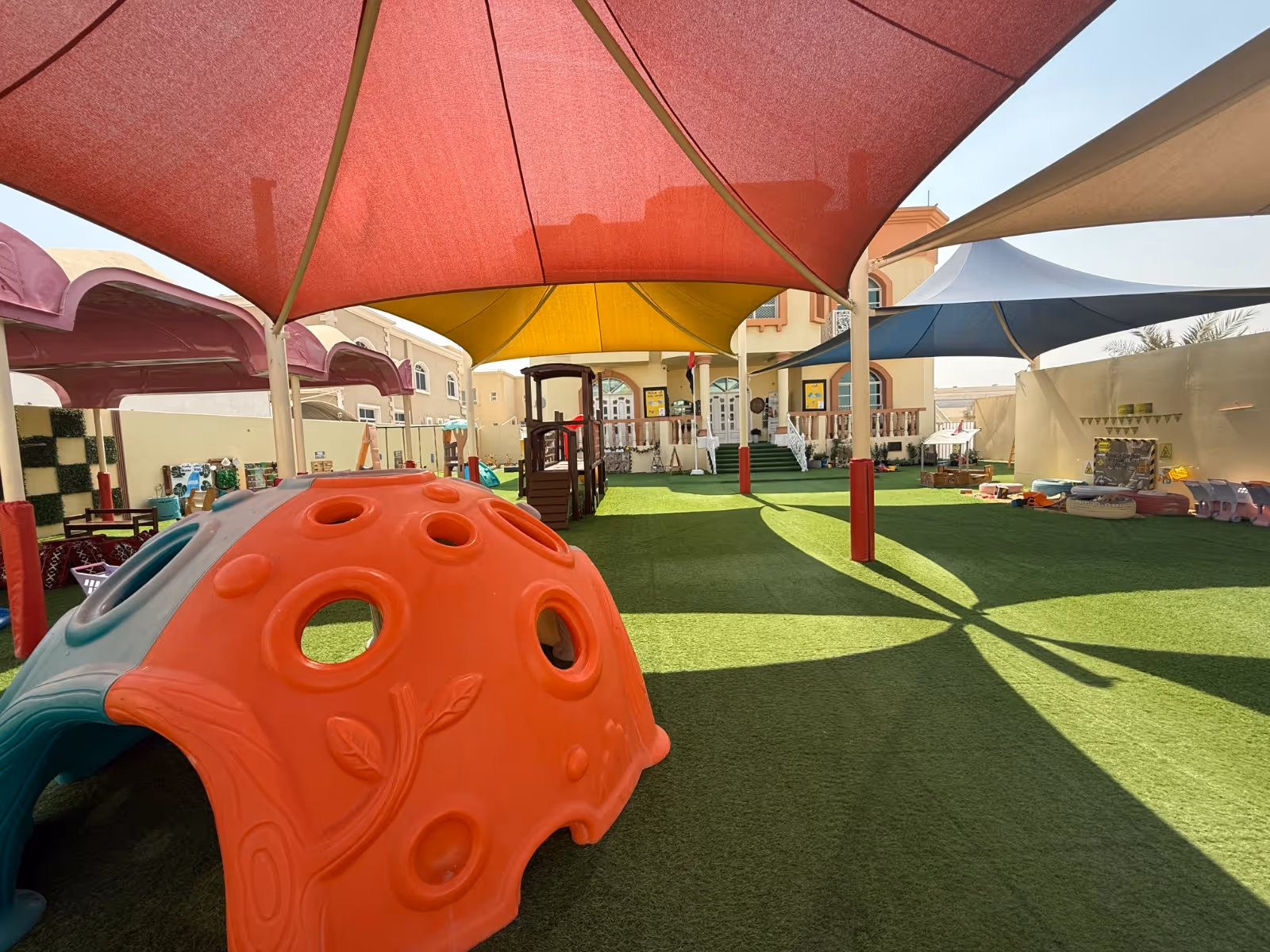 Outdoor children's playground with artificial grass, colorful play structures, and shade sails in red, yellow, and blue.