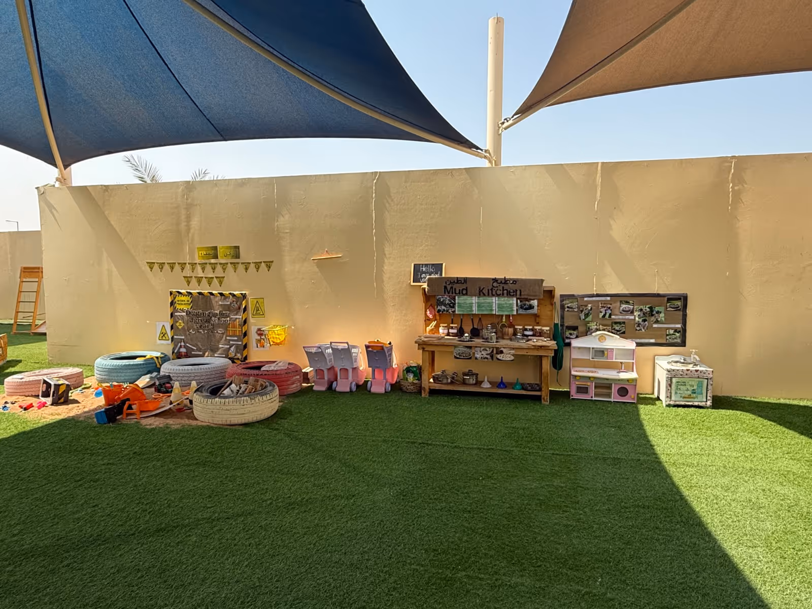 Outdoor children's play area with artificial grass, colorful tire sandbox, toy shopping carts, and a wooden mud kitchen setup under shade sails.