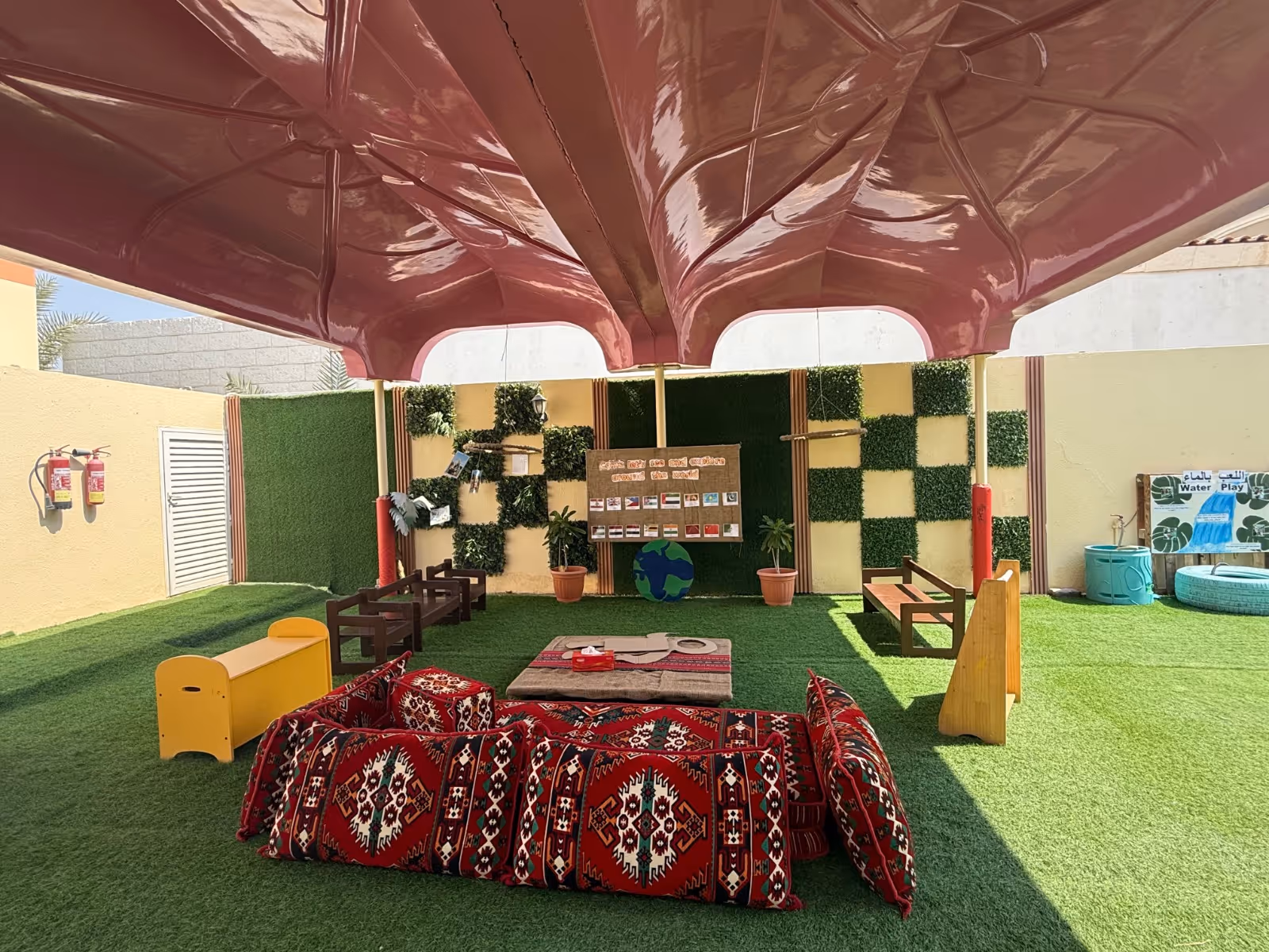 Outdoor shaded seating area with traditional red patterned cushions, wooden benches, artificial grass, and a wall decorated with green and yellow squares and plants.