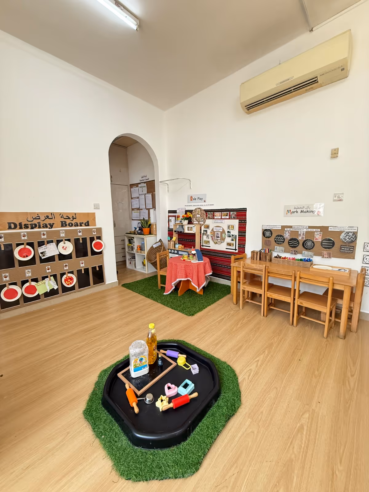 Classroom corner with children's activity areas including a display board, role play area with a table and chair, and a mark making table with small chairs and art supplies.