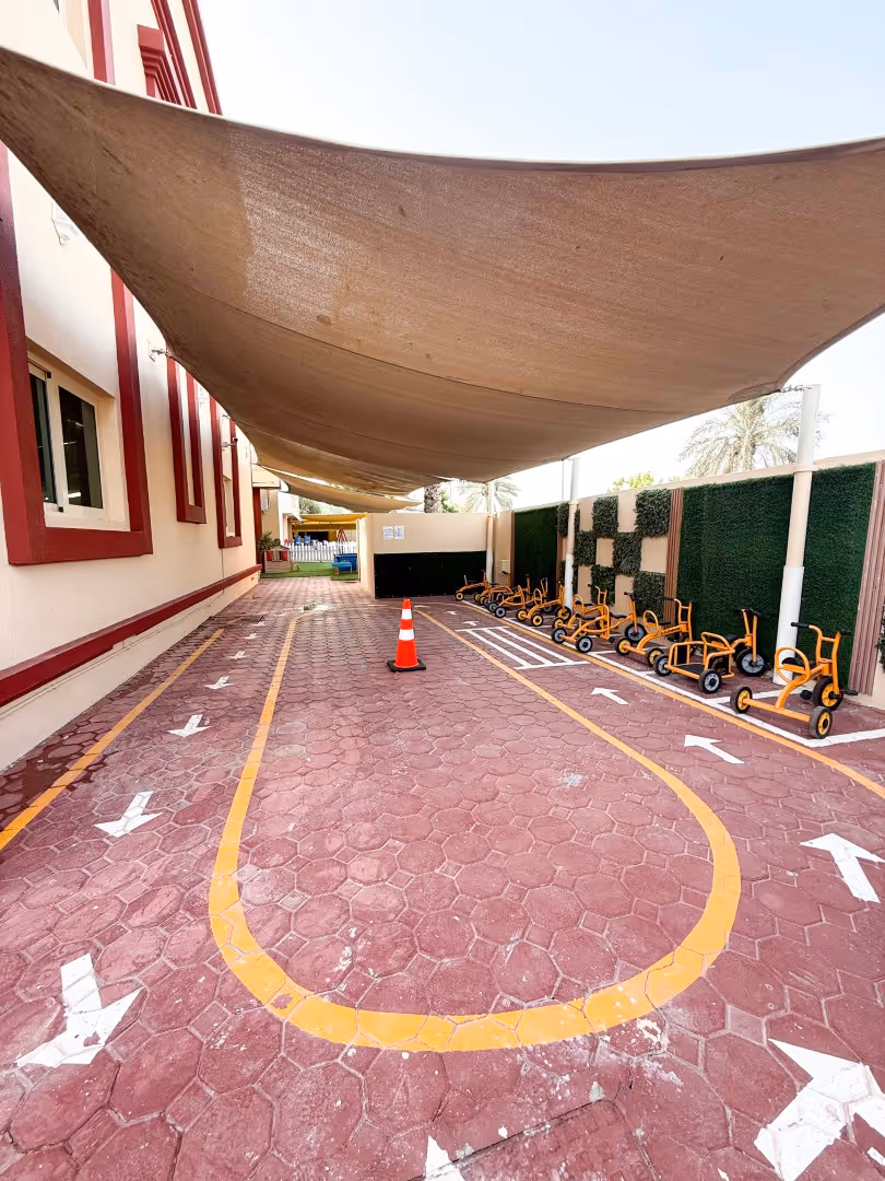 Outdoor play area with a brown shade canopy, a red brick pathway marked with yellow and white arrows, an orange traffic cone, and lined-up yellow tricycles against a wall decorated with green panels.