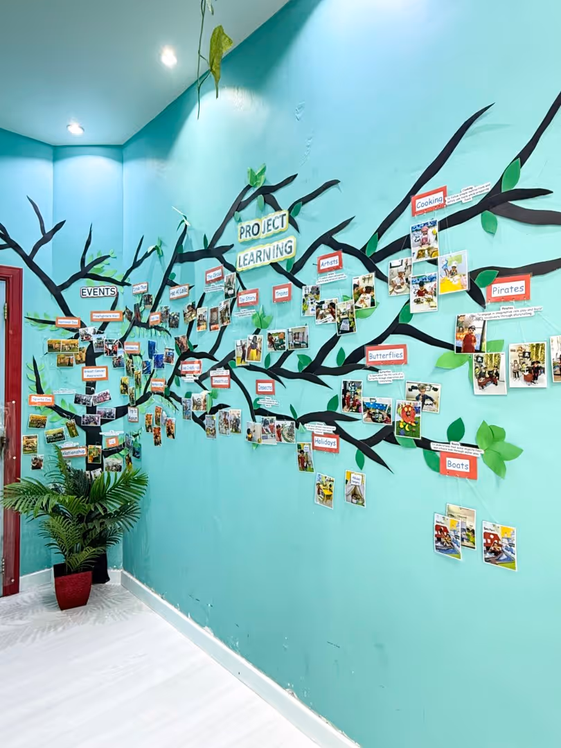 Blue classroom wall decorated with a large black tree mural labeled 'Project Learning,' featuring branches with photo collages and labels such as Events, Artists, Cooking, Pirates, Butterflies, Holidays, and Boats.
