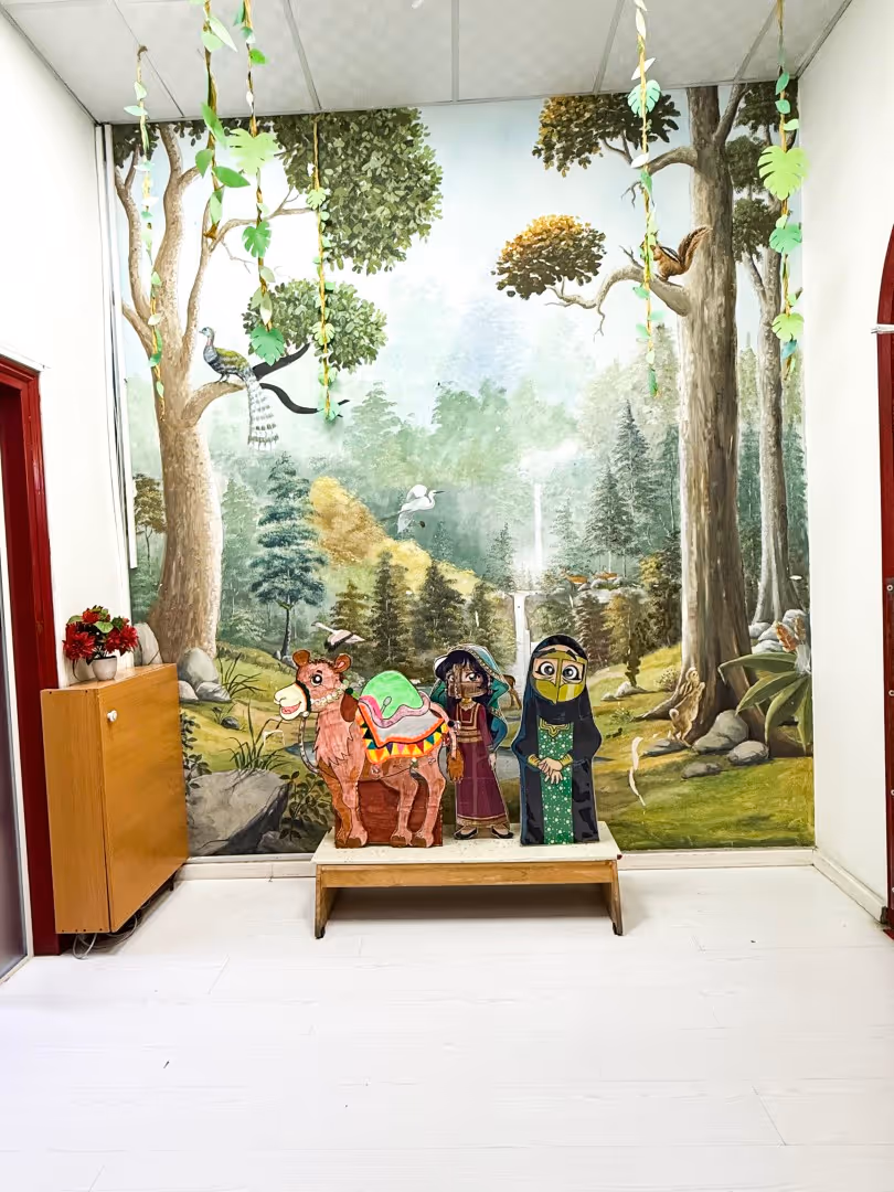 Indoor scene with a colorful painted mural of a forest and birds, featuring cartoon cutouts of a camel, a girl in traditional clothes, and a woman in a green dress with a veil, displayed on a bench.