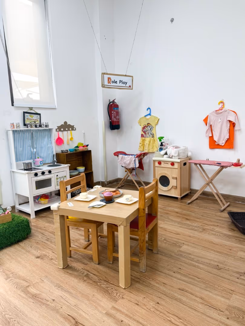 Children's playroom corner with wooden toy kitchen, table set with toy food, and dress-up clothes hanging on the wall labeled Role Play.