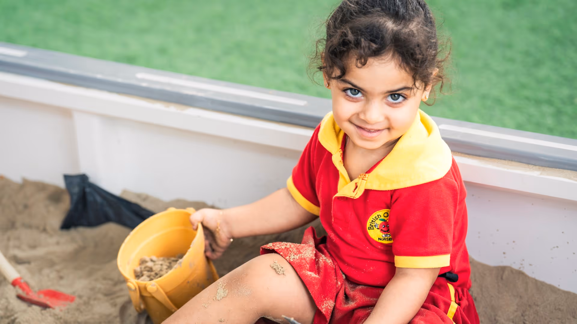 Young girl in a red and yellow nursery uniform playing with sand and a yellow bucket.