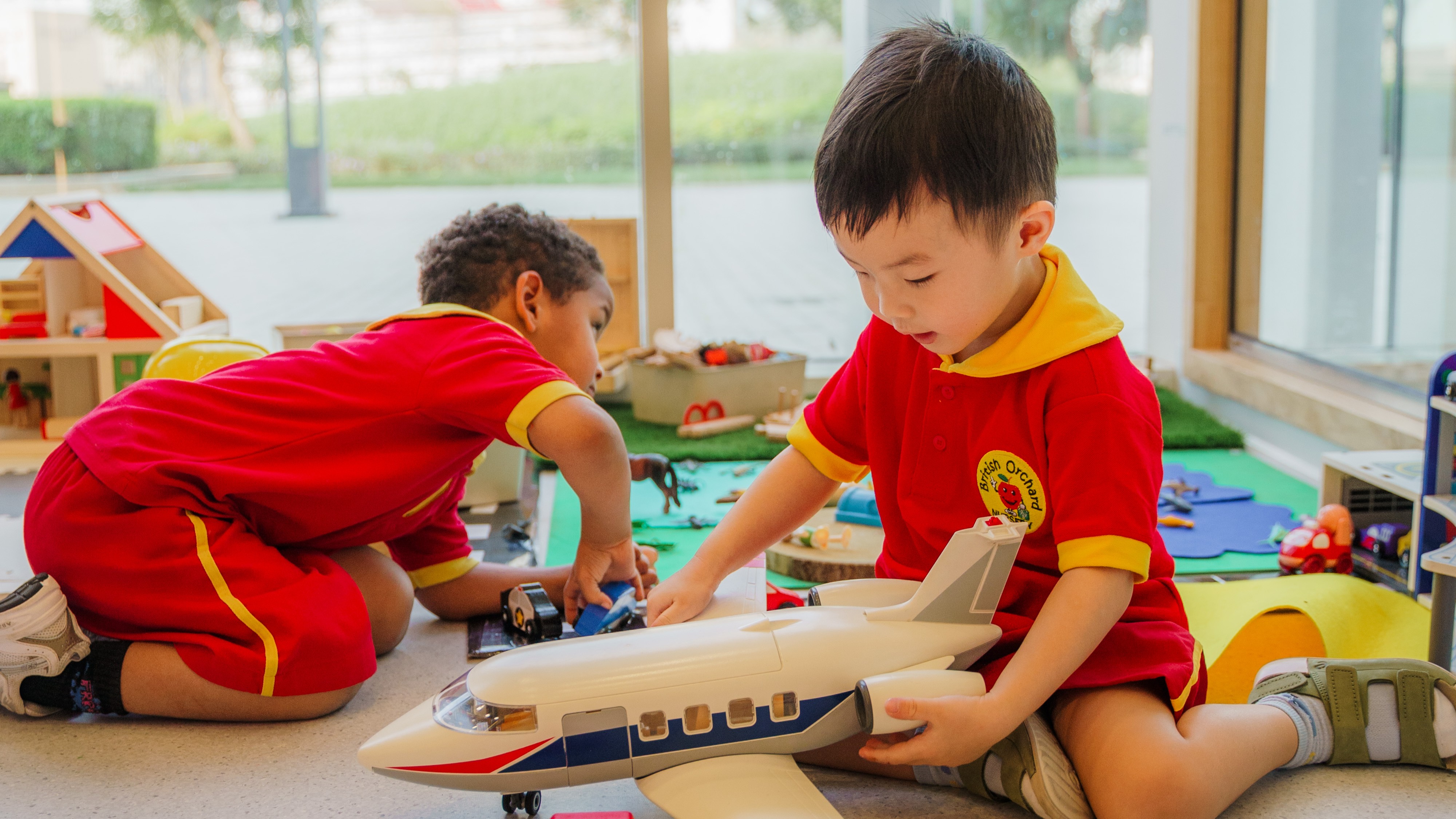 Two young boys in red uniforms playing indoors with toy airplane and cars.
