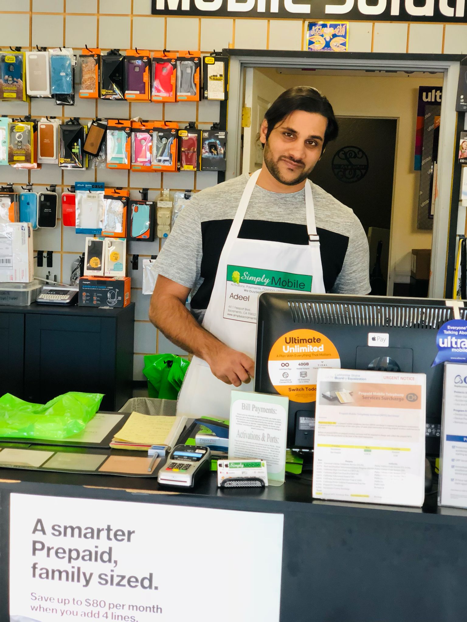 Adeel, owner of Simply Mobile, standing behind the store counter with accessories displayed on the wall and service materials at the register