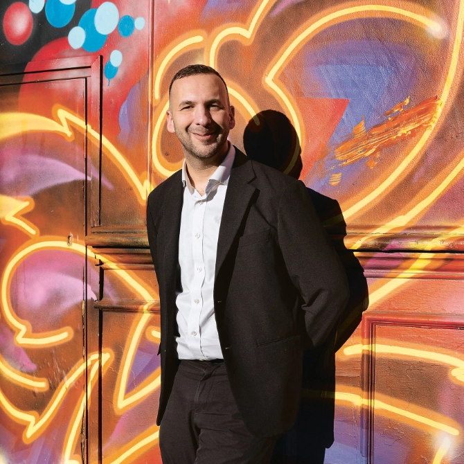 Zack Polanski in schwarzem Anzug lehnt lächelnd vor bunter Wand mit leuchtendem Graffiti